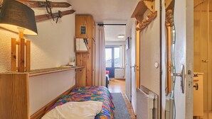 Un couloir étroit mène à une chambre à coucher qui contient un lit, une étagère en bois, une lampe et une fenêtre donnant sur l’extérieur.