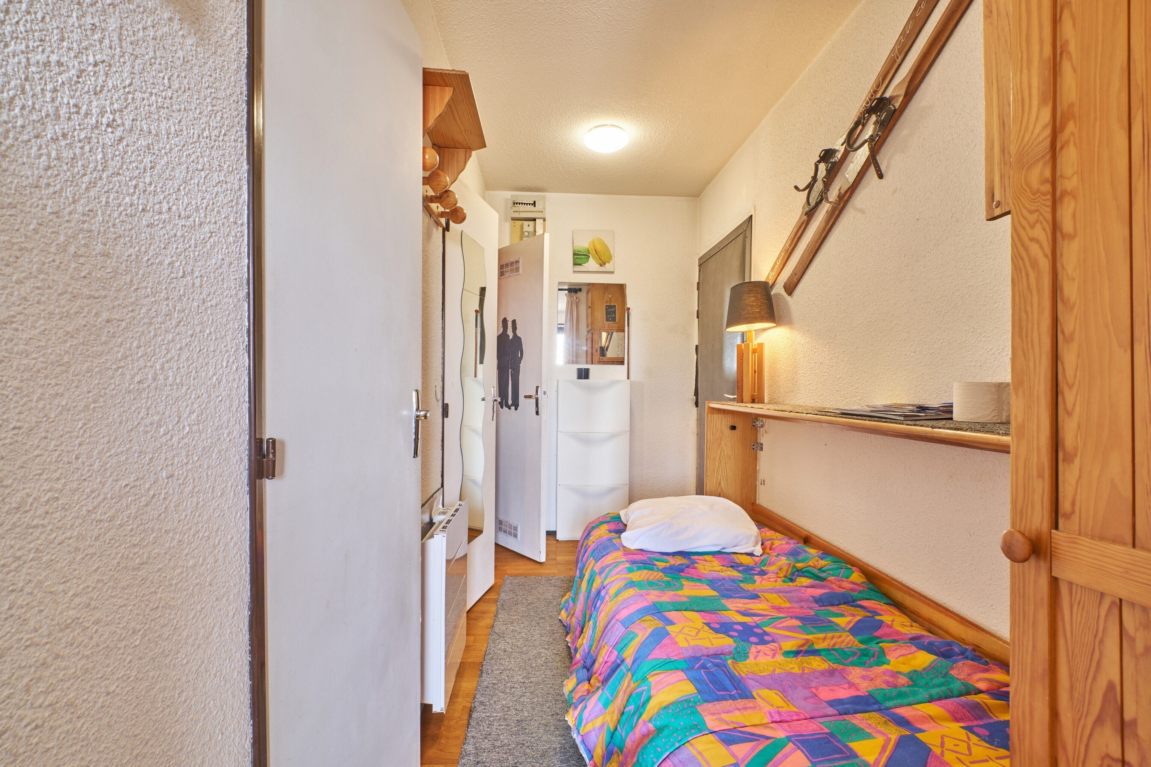 Um corredor estreito que dá para um quarto com edredom acolchoado e colorido, uma estante de madeira com abajur, e uma porta com moldura de madeira.