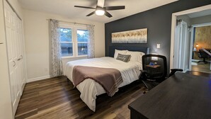 A bedroom with a bed, a ceiling fan, a window with curtains, a painting on the wall, and a desk with a chair.