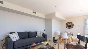 Una sala de estar moderna con un sofá gris, una mesa de centro de vidrio y una zona de comedor con sillas blancas y una mesa de vidrio.