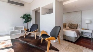 Una habitación de hotel moderna con una cama grande, dos sillas, una planta pequeña y una mesa consola blanca.