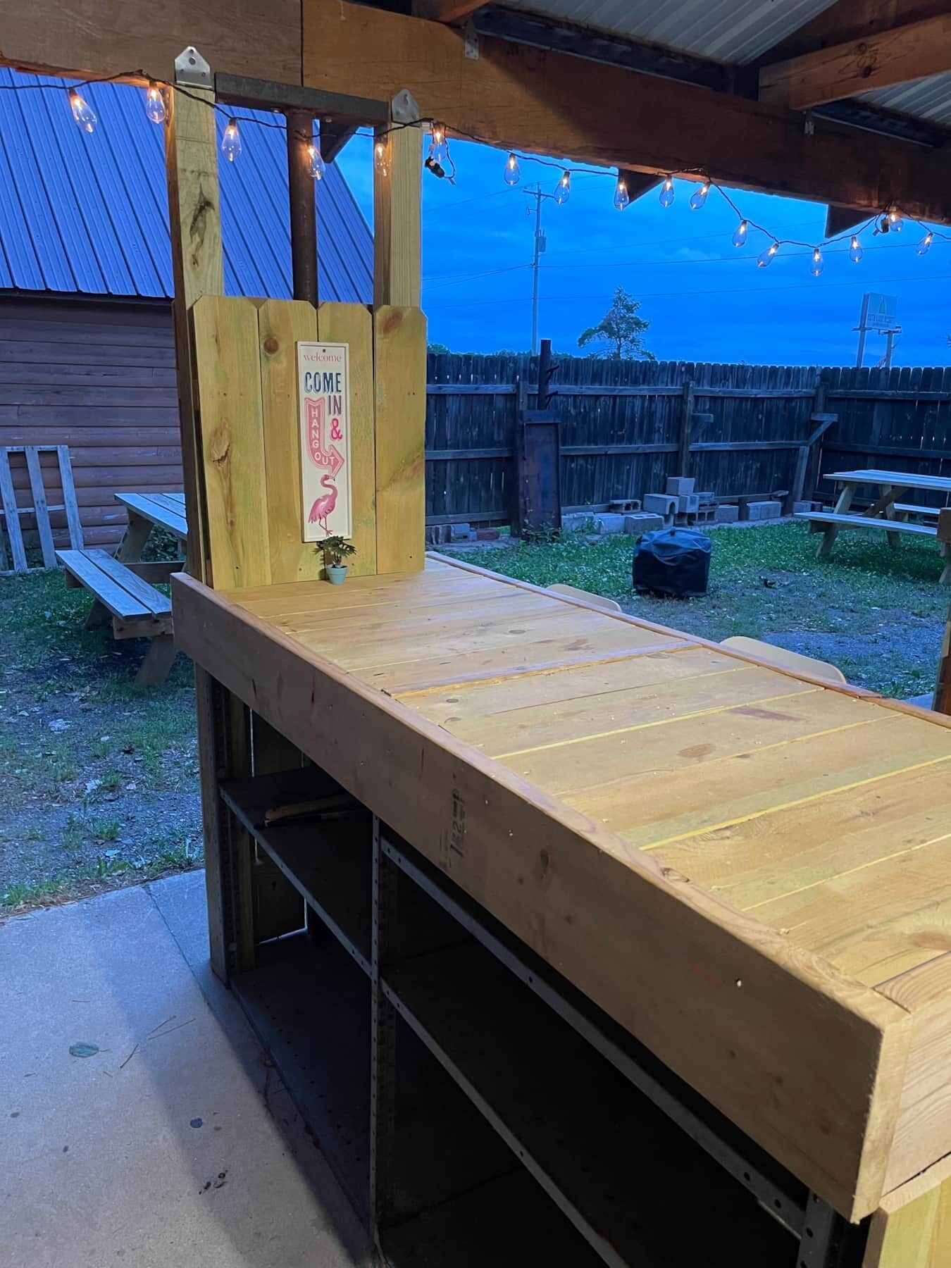 Un mostrador de madera con un letrero que dice 'Welcome Hang in & Out', iluminación exterior y mesas de picnic en un patio trasero.
