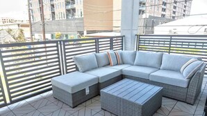 Ein Balkon mit Ecksofa, Ottoman und Fliesenboden, sowie einem Geländer.