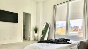 Ein modernes Schlafzimmer mit einem großen Bett, einem Deckenventilator, einem an der Wand montierten Fernseher und Blick auf die Stadt durch große Fenster.