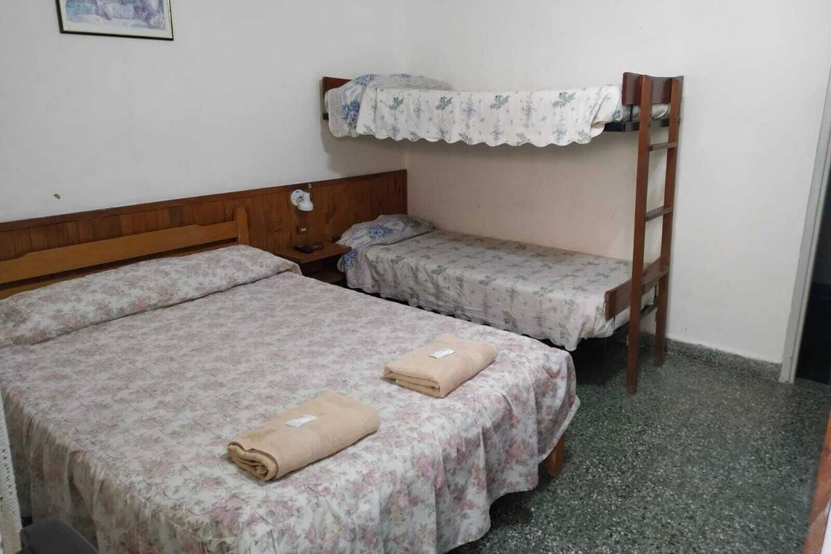 Habitación tipo dormitorio con dos camas, una litera de madera, una mesita de noche pequeña con lámpara y un cuadro en la pared.