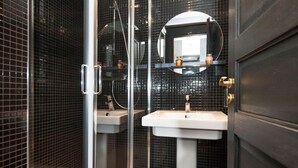 Un baño moderno con paredes de azulejos negros, cabina de ducha de vidrio y dos lavamanos blancos.