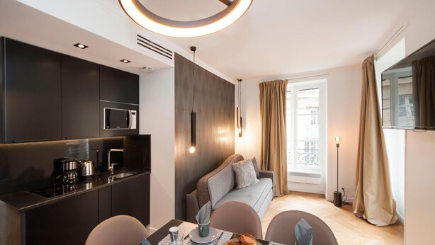 Una habitación de hotel moderna con kitchenette, comedor, sofá y un gran ventanal con cortinas.