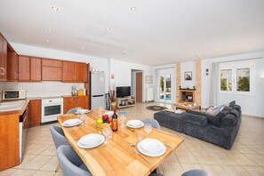 A modern kitchen with wooden cabinets, a dining table set for four, and a living area with a sofa and TV.