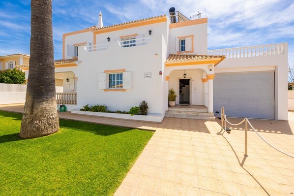 Une maison à deux étages, avec une façade blanche, des bordures orange et un toit recouvert de tuiles. Un garage se trouve sur la droite, et une petite véranda avec des marches mène à l’entrée.