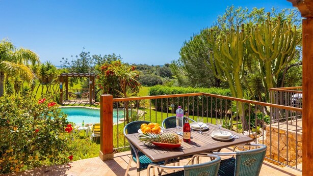 Une terrasse en bois avec une table dressée pour le repas, donnant sur une piscine et un jardin luxuriant.