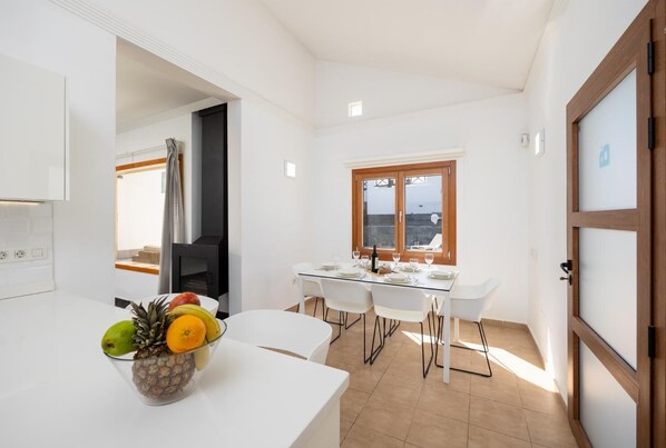 A modern kitchen with white cabinets, a dining area with a glass table, and a view through a window. - Villa Dacil - Three Bedroom Villa, Sleeps 6 (Playa Blanca)