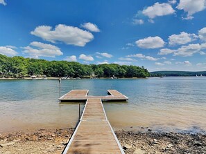 A wooden dock extending into a calm lake with a forested shoreline and a clear sky. - Lake Shore by Taylor Made Deep Creek (Swanton)