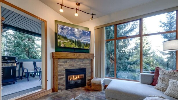 A spacious living room with a fireplace, a large window with a view of trees, and a cozy seating area. - Central 2BR with Prime Amenities (Whistler)