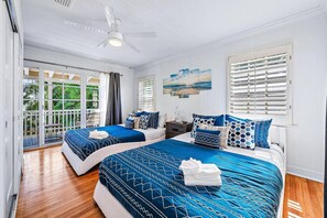 A hotel room with two beds, a ceiling fan, a view of greenery, and a painting on the wall.