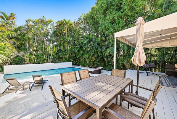 A poolside area with a wooden table and chairs, a covered seating area, and a pool.