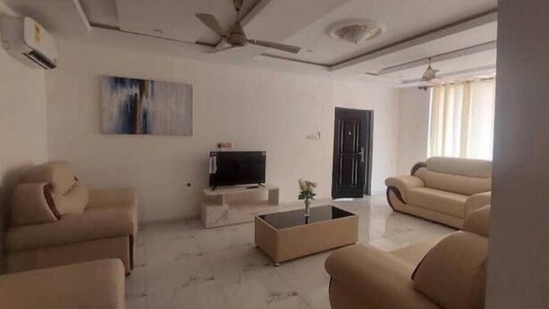 A modern living room with a flat-screen TV, beige sofas, a coffee table, and a ceiling fan. - Luxury complex at Agric Kumasi (Kumasi)