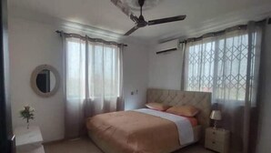 A bedroom with a bed, a ceiling fan, a nightstand, a lamp, and a window with curtains. - Luxury complex at Agric Kumasi (Kumasi)