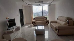 A modern living room with a large window, a flat-screen TV, and two beige sofas. - Luxury complex at Agric Kumasi (Kumasi)