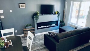 A modern living room with a grey sofa, a dining table with chairs, a flat-screen TV mounted above a fireplace, and a large window providing natural light. - The Cottages @Gull&Castle #2
LOCATION, LOCATION, LOCATION!
Downtown Wasagaming

 (Wasagaming)