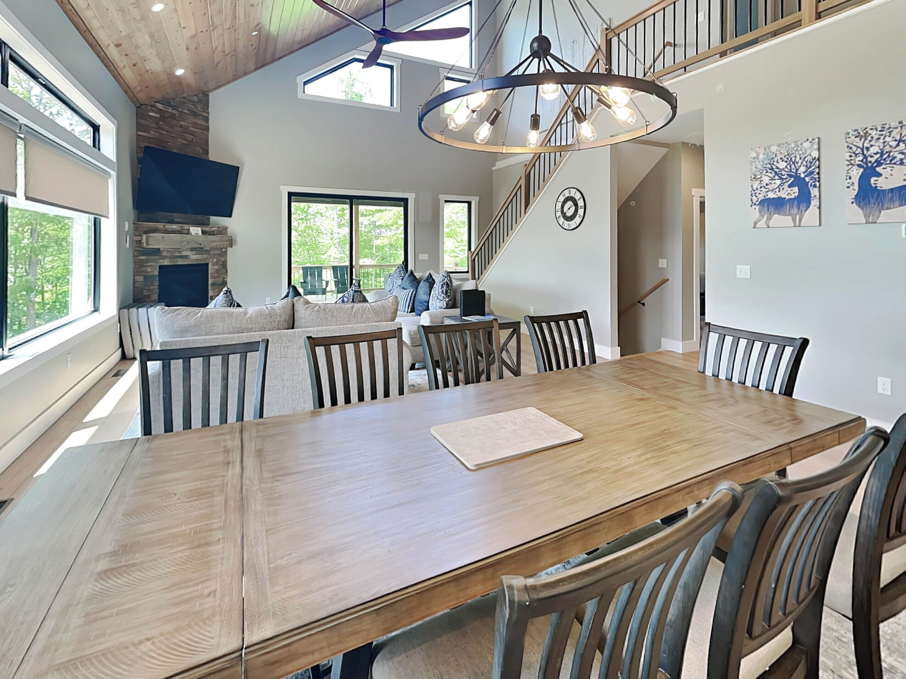 A spacious dining area with a large wooden table and chairs, a modern chandelier, and a staircase leading to an upper level.