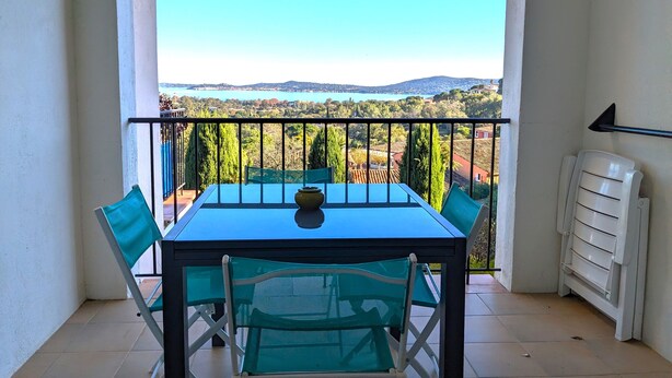 A balcony with a table and chairs, offering a view of a lake and hills. - Superbe Vue Mer 2 Pièces Climatisé Les Restanques (Grimaud)
