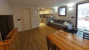 A modern apartment with a wooden dining table, grey sofa, and a kitchenette in the background.