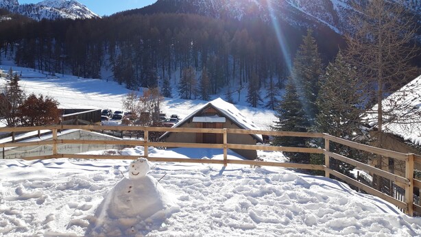 A snowman in a snowy area with a wooden fence, a cabin, and a mountain in the background.