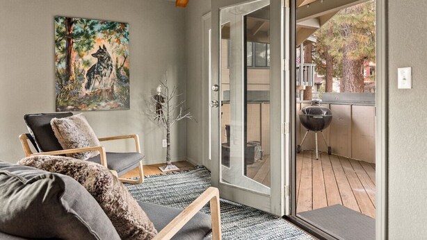 A cozy living room with a sofa, armchair, and a painting of a dog. A glass door leads to an outdoor deck with a grill. - Moonridge 2BR Ski Oasis (Big Bear Lake)