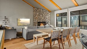 A modern living room with a stone wall fireplace, a grey sofa, a wooden dining table with chairs, and large windows. - Moonridge 2BR Ski Oasis (Big Bear Lake)