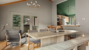A modern kitchen with a large island, wooden chairs, and a bench. There is a dining table with chairs, a chandelier, and a view of the outdoors through a window. - Moonridge 2BR Ski Oasis (Big Bear Lake)