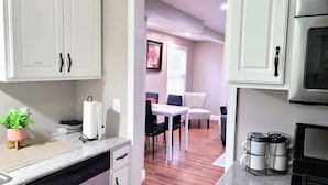 A kitchen with white cabinets, a dishwasher, a countertop with a plant, a coffee maker, and a dining area with a table and chairs.