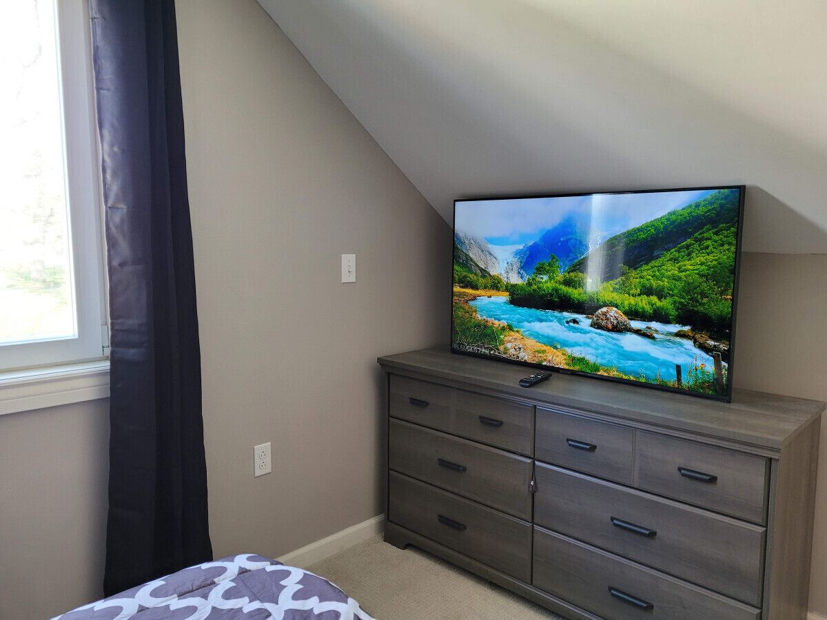 Un dormitorio con televisor sobre un tocador, una ventana con cortinas y una cama con colcha estampada.