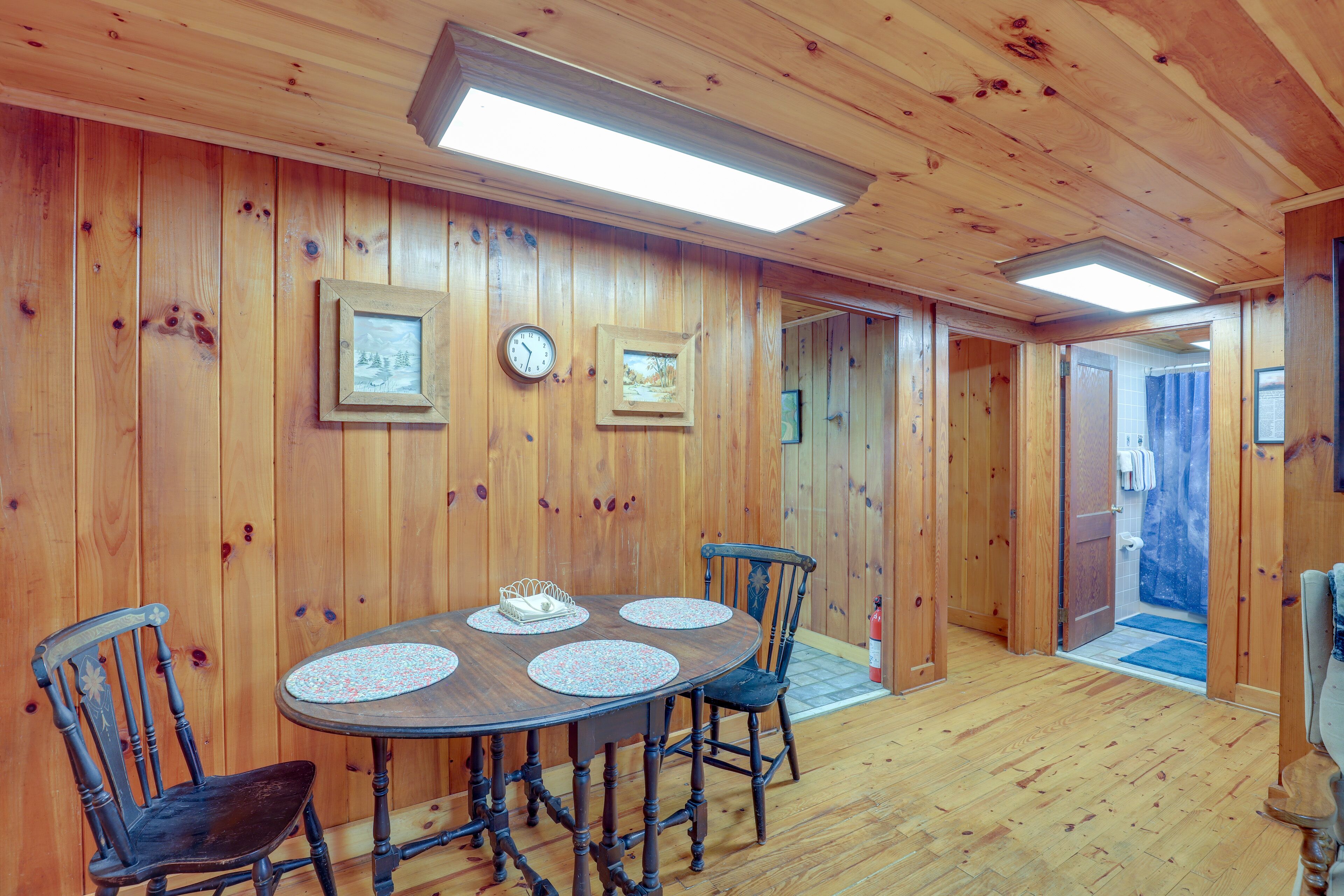A wooden-paneled room with a round table and four chairs, a clock, and framed pictures on the walls.