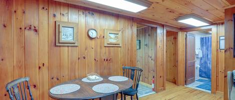 A wooden-paneled room with a round table and four chairs, a clock, and framed pictures on the walls.