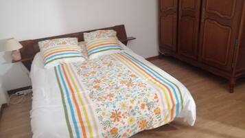 A bed with a floral bedspread and striped pillows, a wooden headboard, a bedside table with a lamp, and a wooden wardrobe.