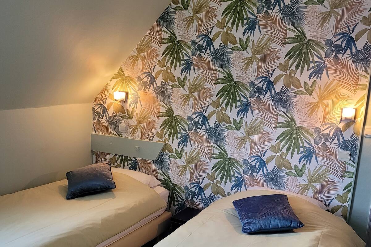 Une chambre avec deux lits simples, chacun équipé d’un oreiller et d’une table de chevet. Les murs sont décorés d’un motif de feuilles tropicales.