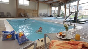 An indoor swimming pool with lounge chairs, a poolside table with a tray of food and drinks, and a child swimming. - Ferme du Château (Monampteuil)