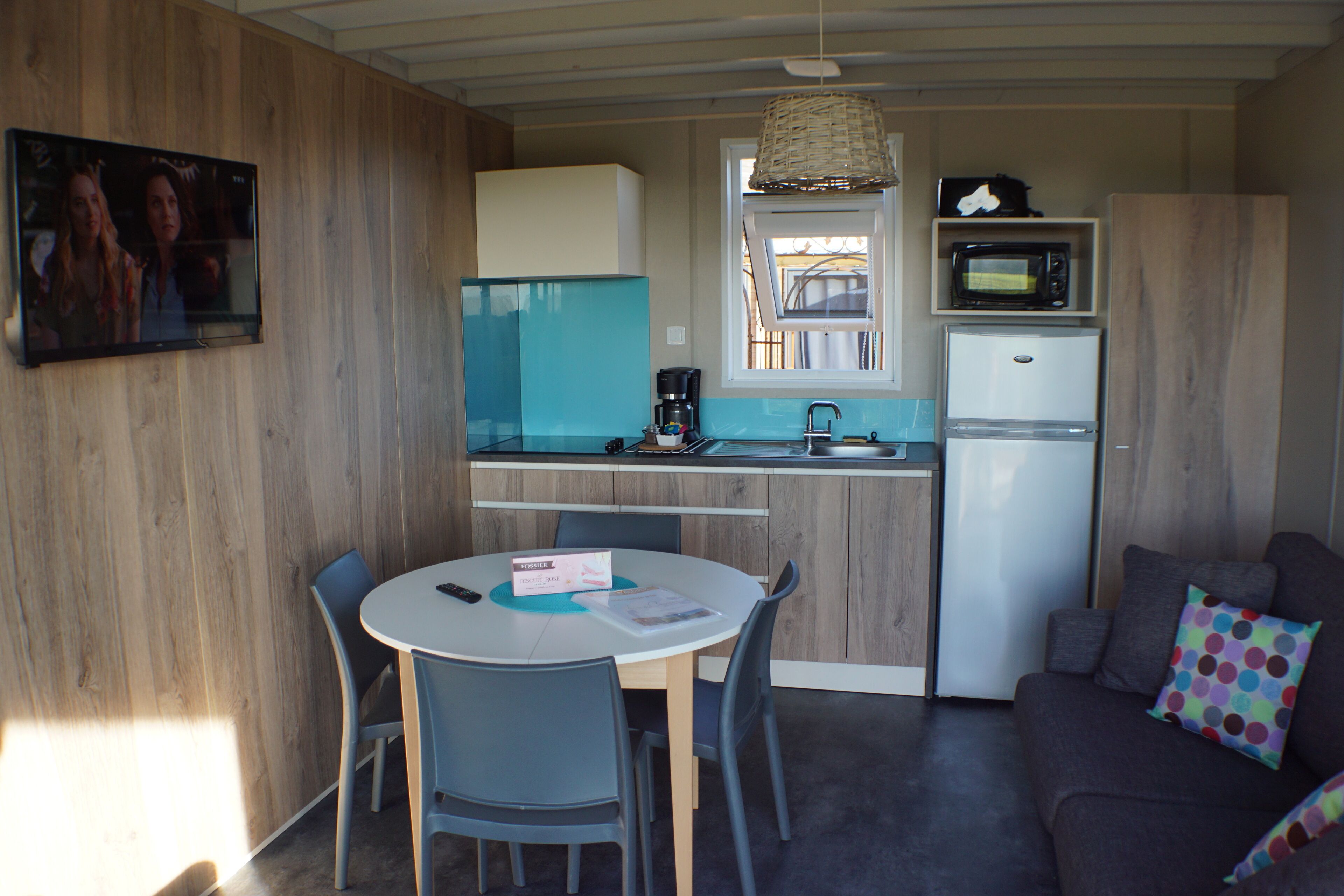 Cocina compacta con microondas, refrigerador y cafetera en la encimera. Hay una mesa de comedor con sillas y un televisor montado en la pared.