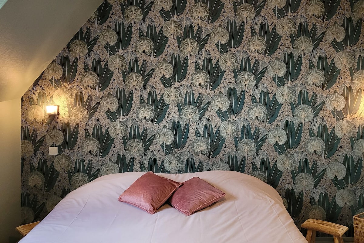 Une chambre à coucher avec un lit, des oreillers, une serviette pliée, un petit tabouret en bois et un mur orné d’un motif de feuilles.