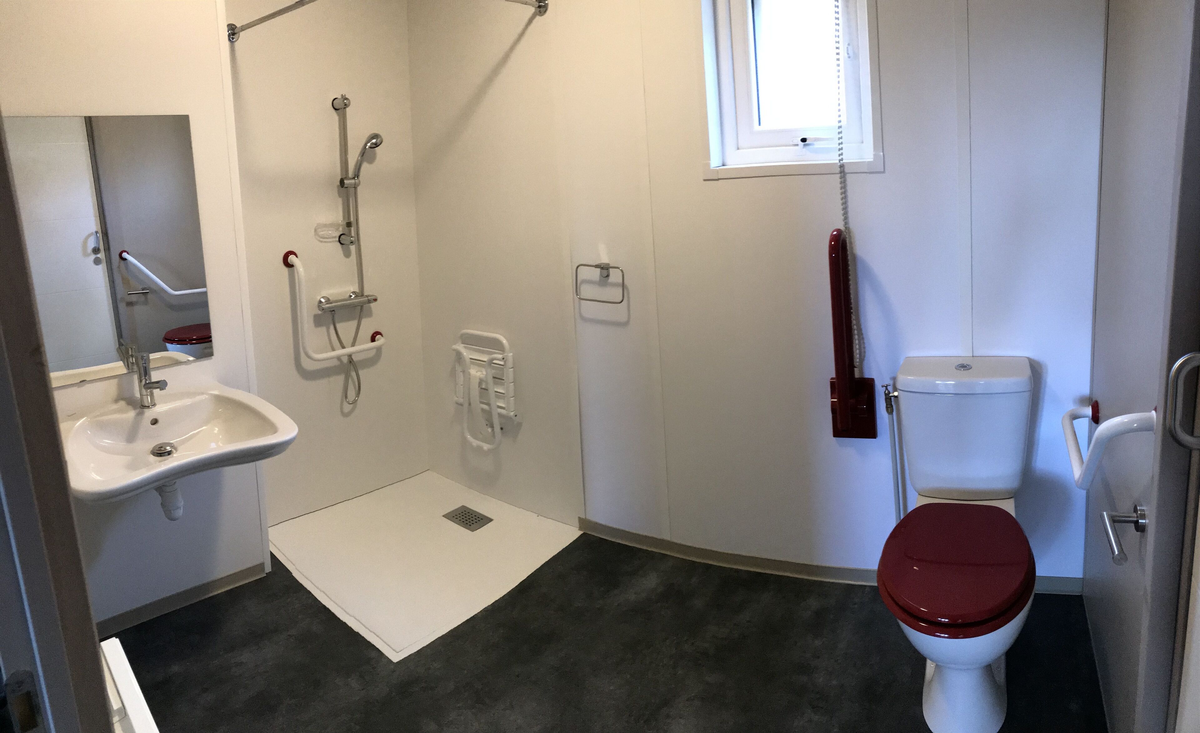 Une salle de bain avec un lavabo blanc, une douche équipée d’une barre d’appui, des toilettes avec un abattant rouge et une fenêtre avec des stores.