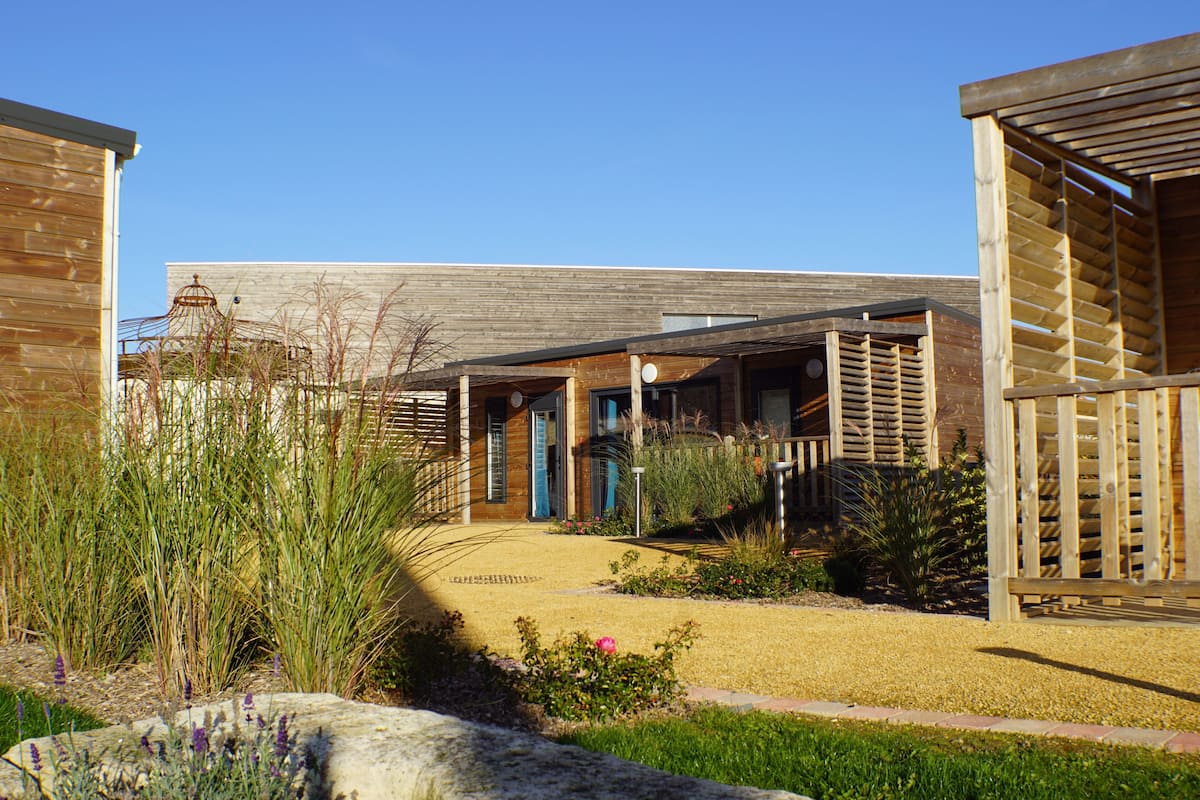 Un bâtiment moderne doté d’un patio couvert, d’un chemin menant à l’entrée et d’un jardin bien entretenu où poussent diverses plantes et fleurs.