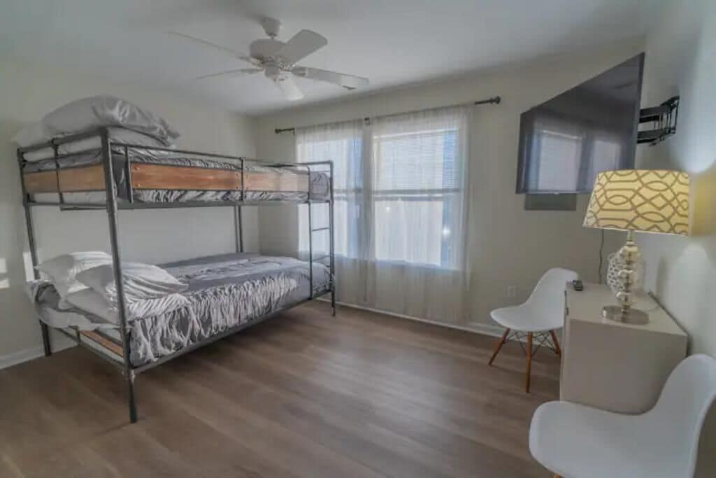 A room with a bunk bed, a desk with a lamp, a chair, and a ceiling fan.