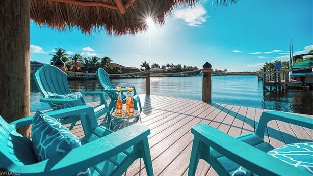 Een houten terras met blauwe stoelen en een tafel met drankjes, met uitzicht op een rustig wateroppervlak en palmbomen.