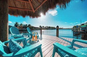 A wooden deck with blue chairs and a table with drinks, overlooking a calm body of water and palm trees.