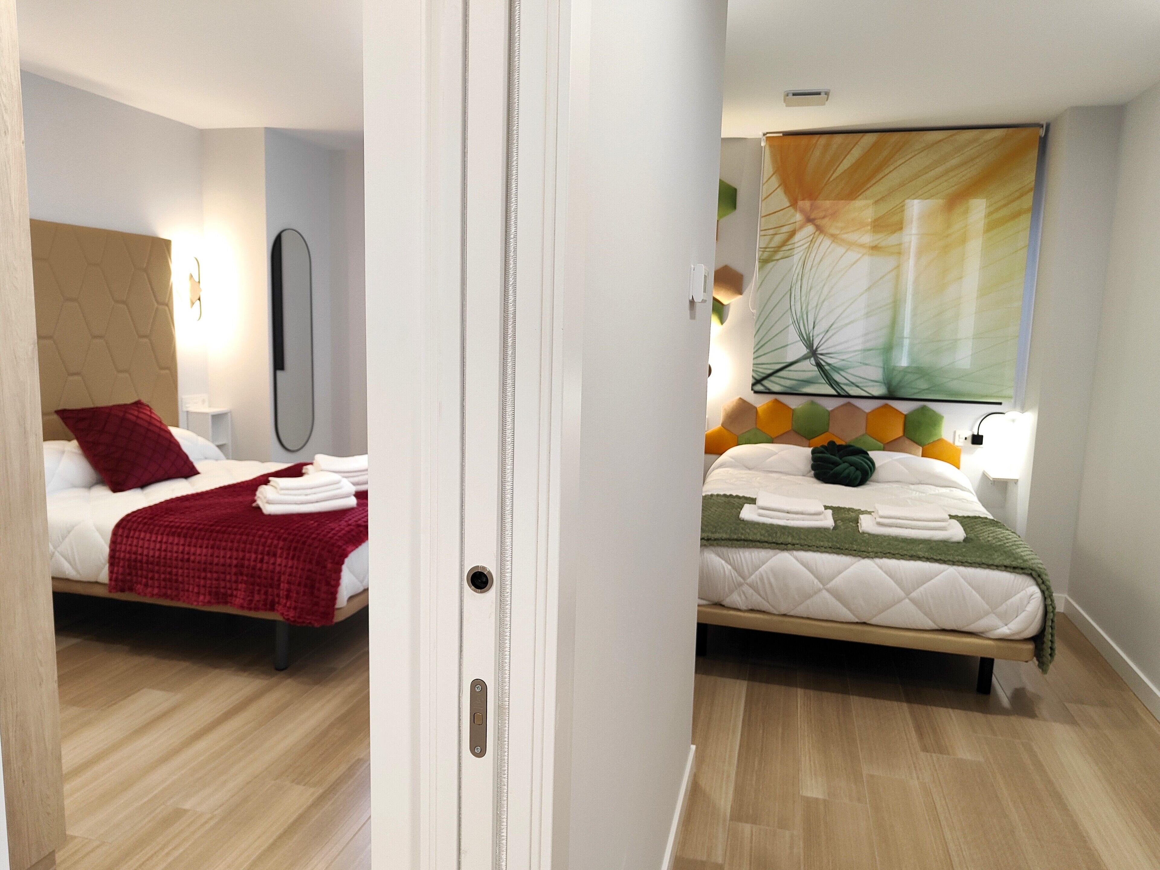 A hotel room with a bed, a mirror, a wall-mounted light, and a window with a plant.