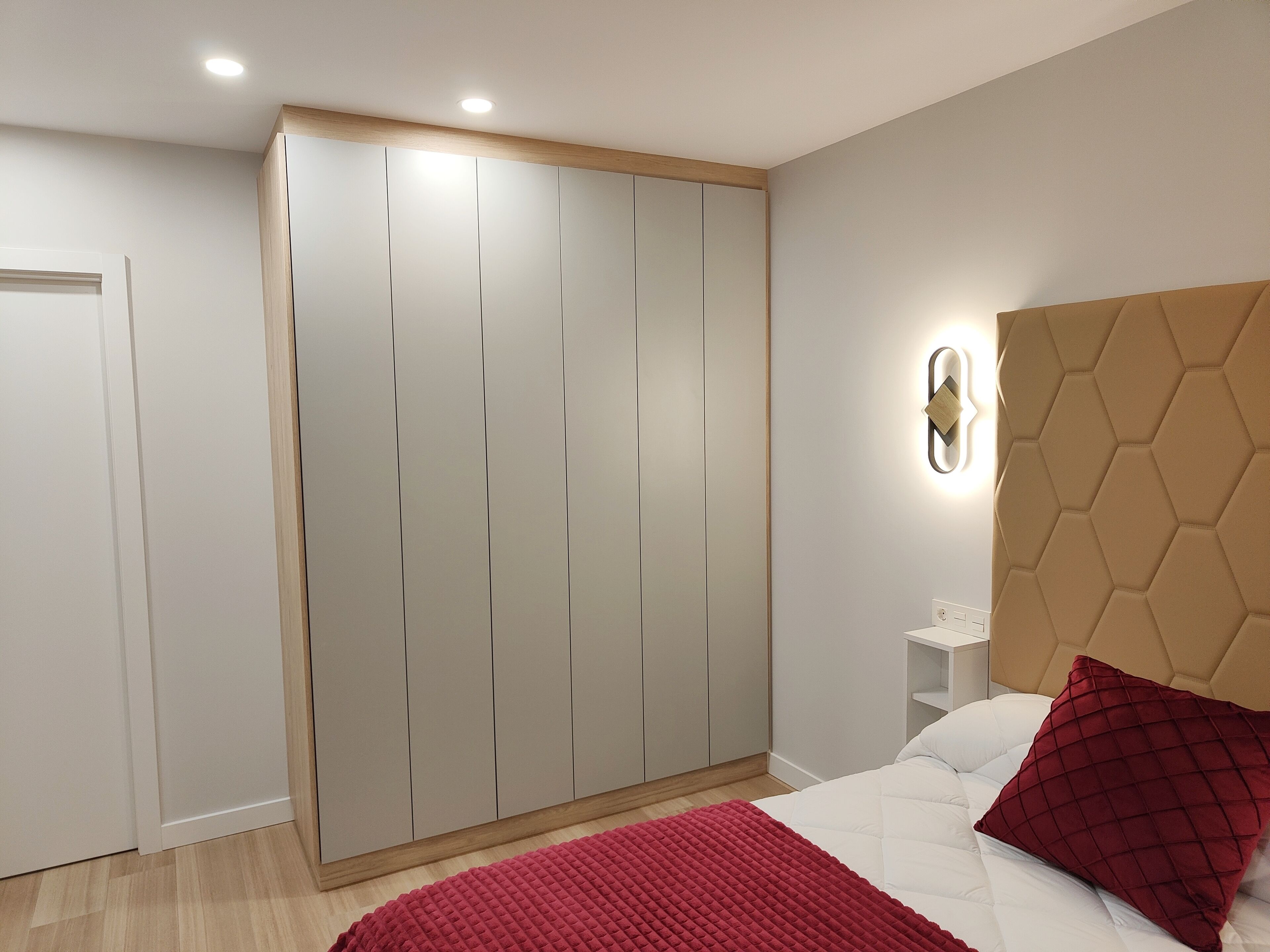 Une chambre à coucher avec un lit, une couverture rouge, une tête de lit, une armoire et un luminaire fixé au mur.