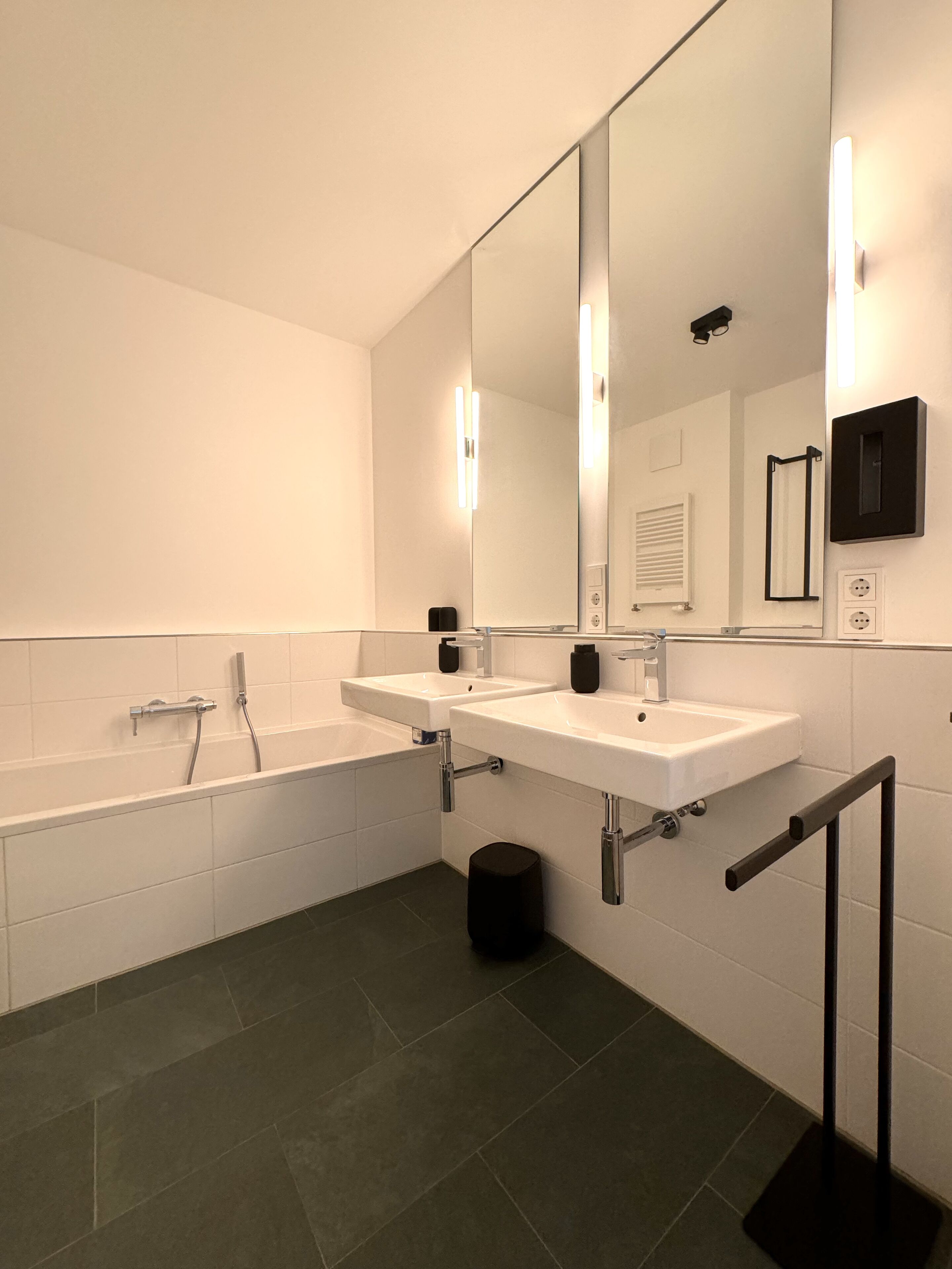 A modern bathroom with a bathtub, two sinks, a large mirror, and a black trash bin.