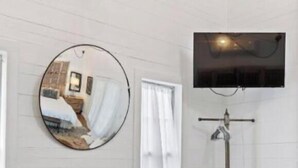 Una habitación con un escritorio de madera, taburetes, una silla, un espejo redondo y un televisor montado en la pared.
