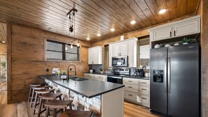 A modern kitchen with wooden walls, a central island with stools, stainless steel appliances, and a large refrigerator.