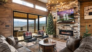 A cozy living room with a stone fireplace, a Christmas tree, leather sofas, and a large window offering a mountain view.